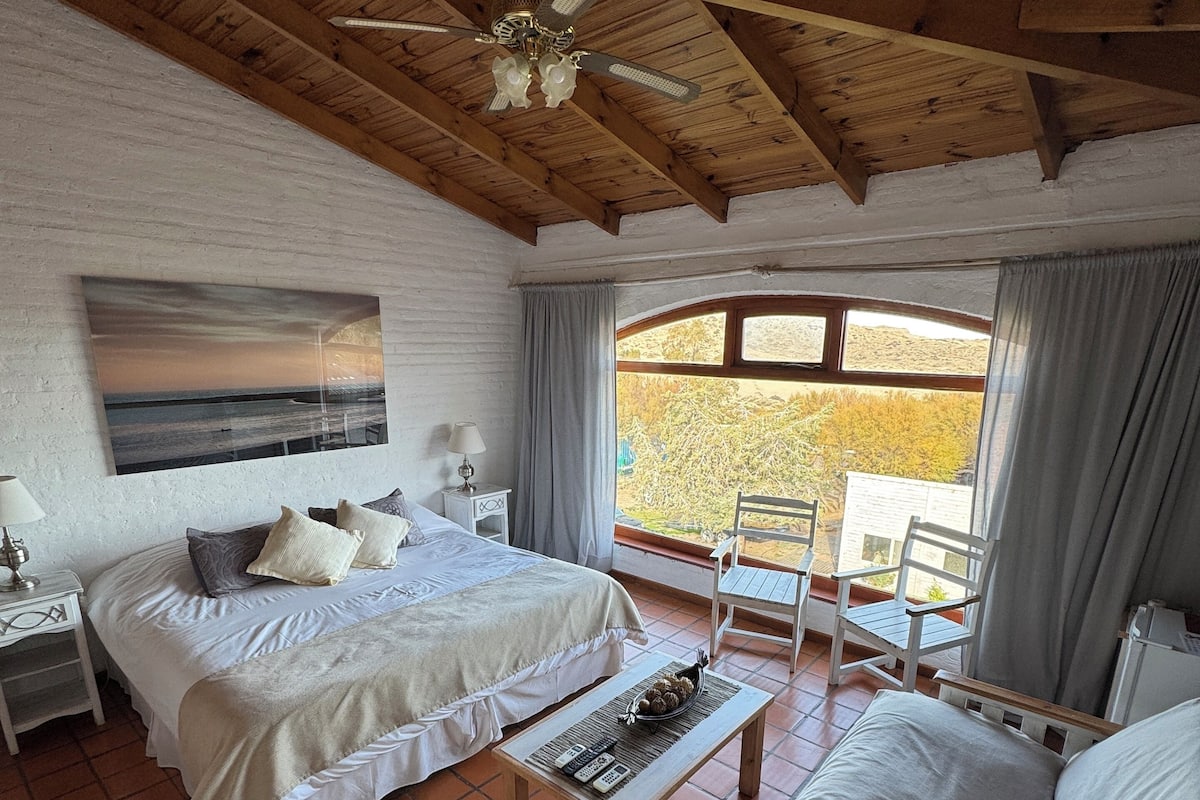 Une chambre à coucher comprenant un lit, un canapé, une table avec des télécommandes, un ventilateur de plafond et une fenêtre avec des rideaux.