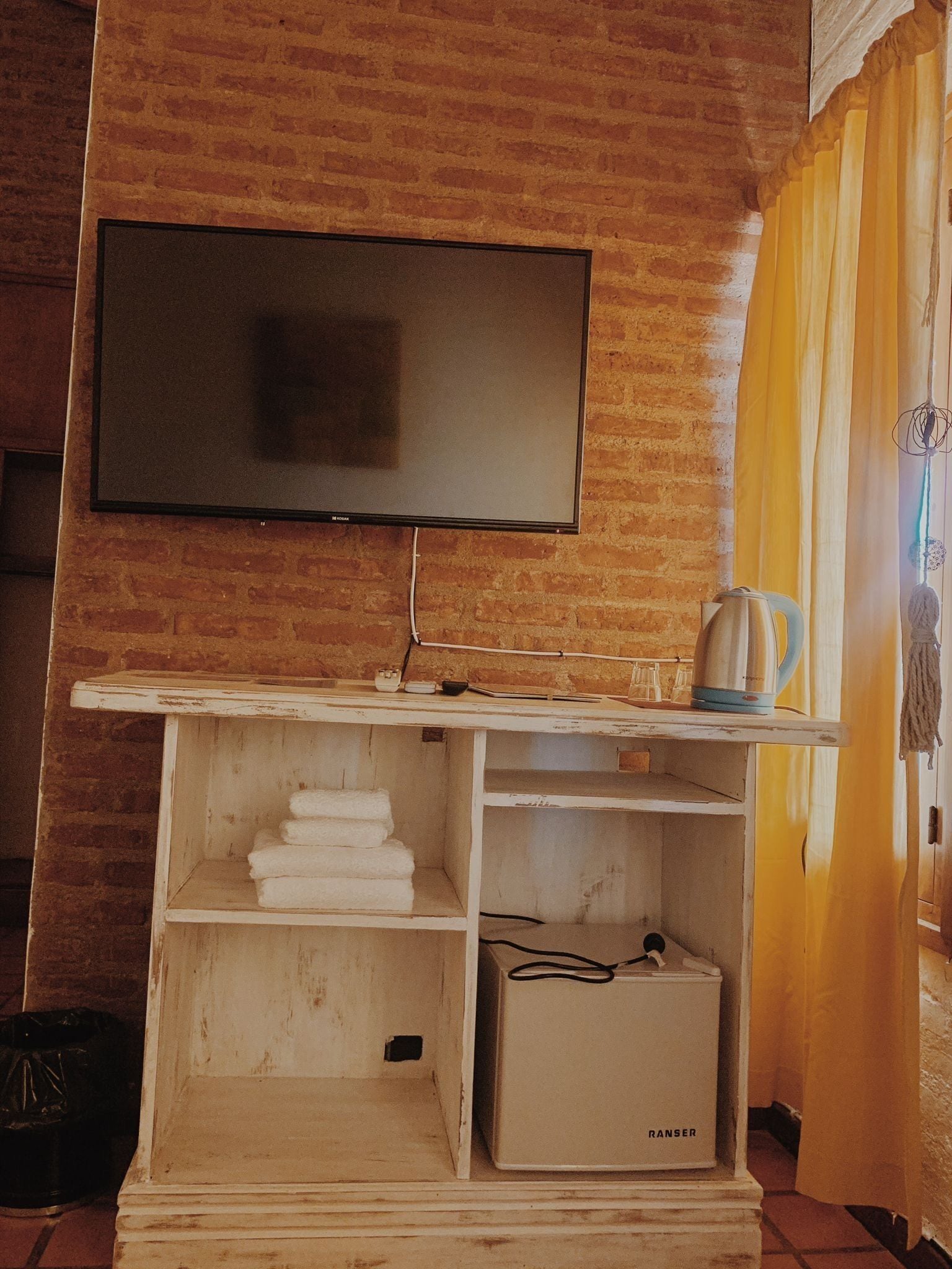 Een kamer met een televisie aan een bakstenen muur, een wit kastje met een RANSEK apparaat, en een geel gordijn.