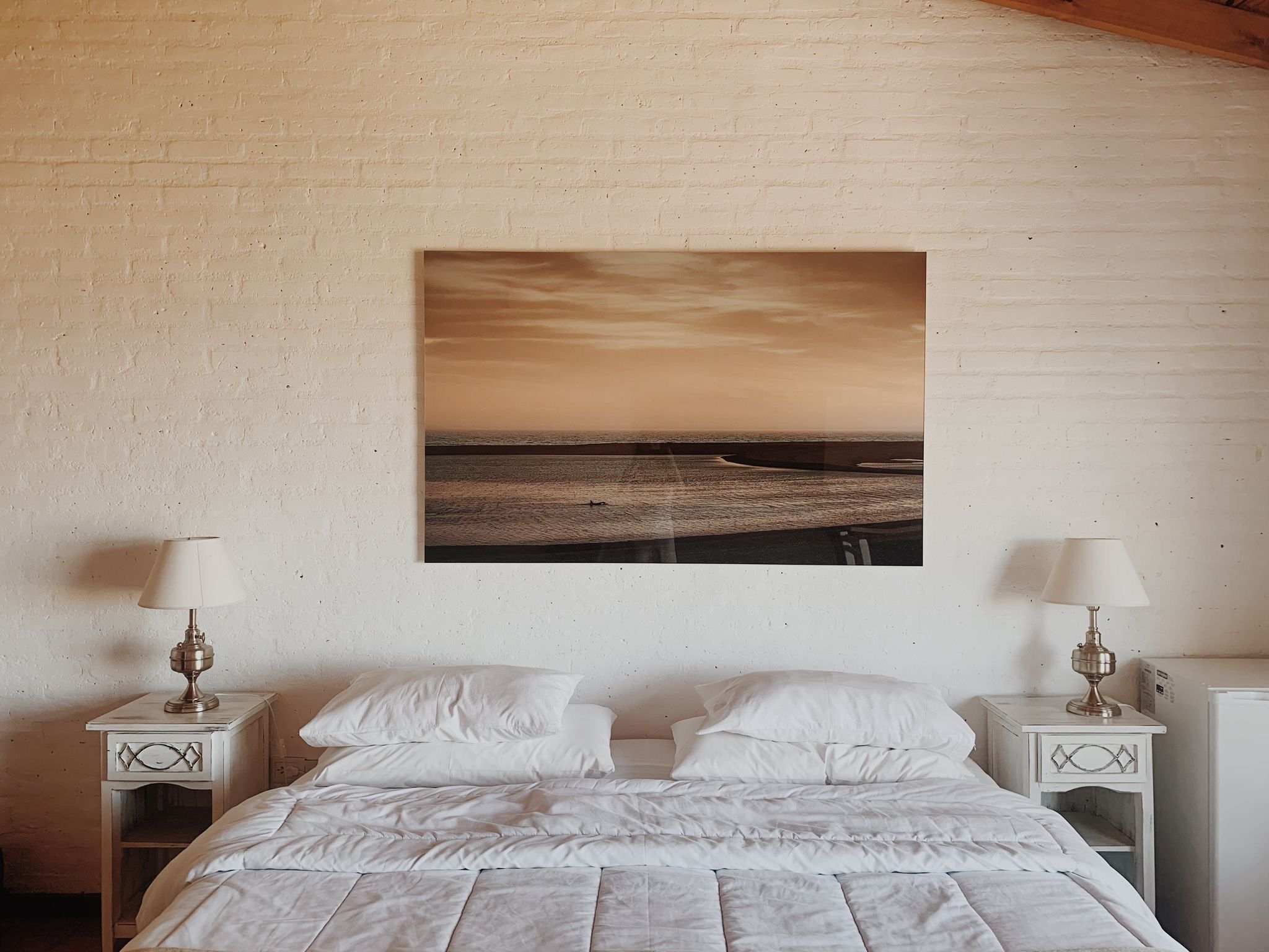 A bedroom with a bed, white bedding, two bedside tables with lamps, and a large landscape painting on the wall.