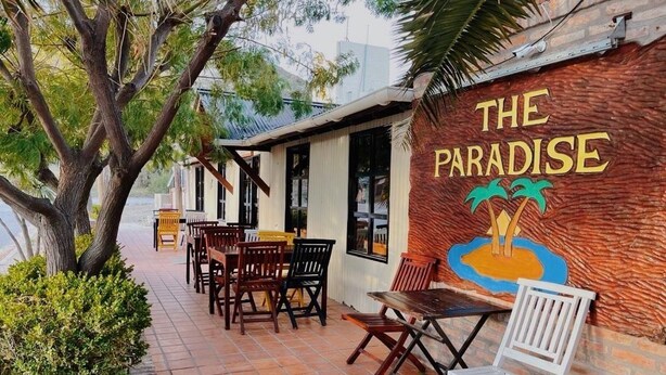 An outdoor seating area with tables and chairs, a mural with palm trees, and a building with a sign reading "THE PARADISE". - The Paradise (Puerto Piramides)