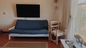 A living room with a white sofa, a television mounted on the wall, a wooden chair, and a small table with a decorative item. - The Paradise (Puerto Piramides)