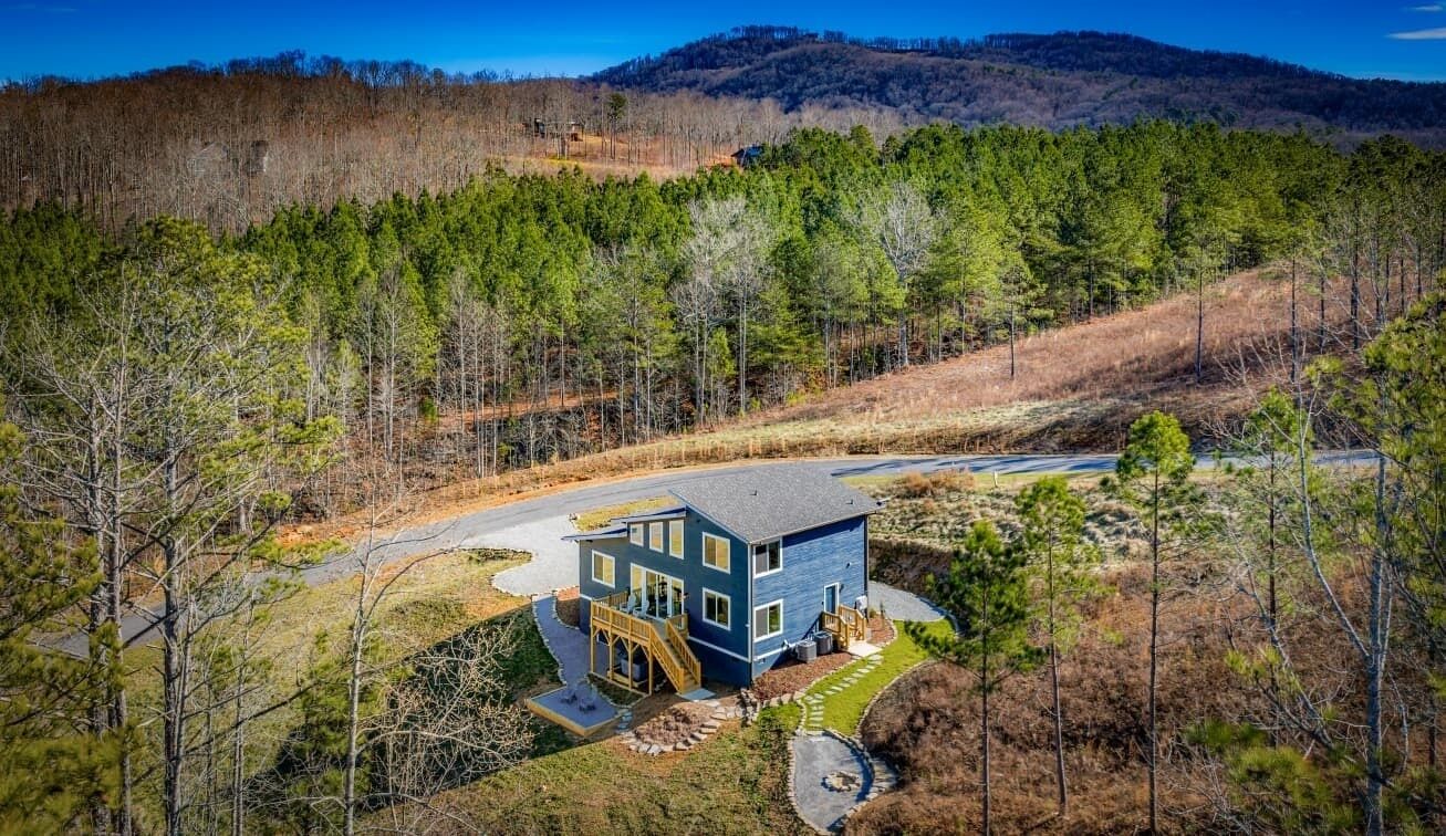 Une maison à deux étages, à l’extérieur bleu, entourée d’une zone boisée sous un ciel dégagé.
