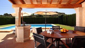 Um pátio externo com mesa e cadeiras de madeira, e piscina ao fundo.