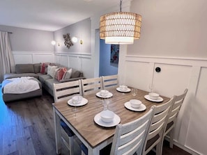 Uma área de refeições com mesa de madeira posta para quatro pessoas, cadeiras brancas e um lustre.