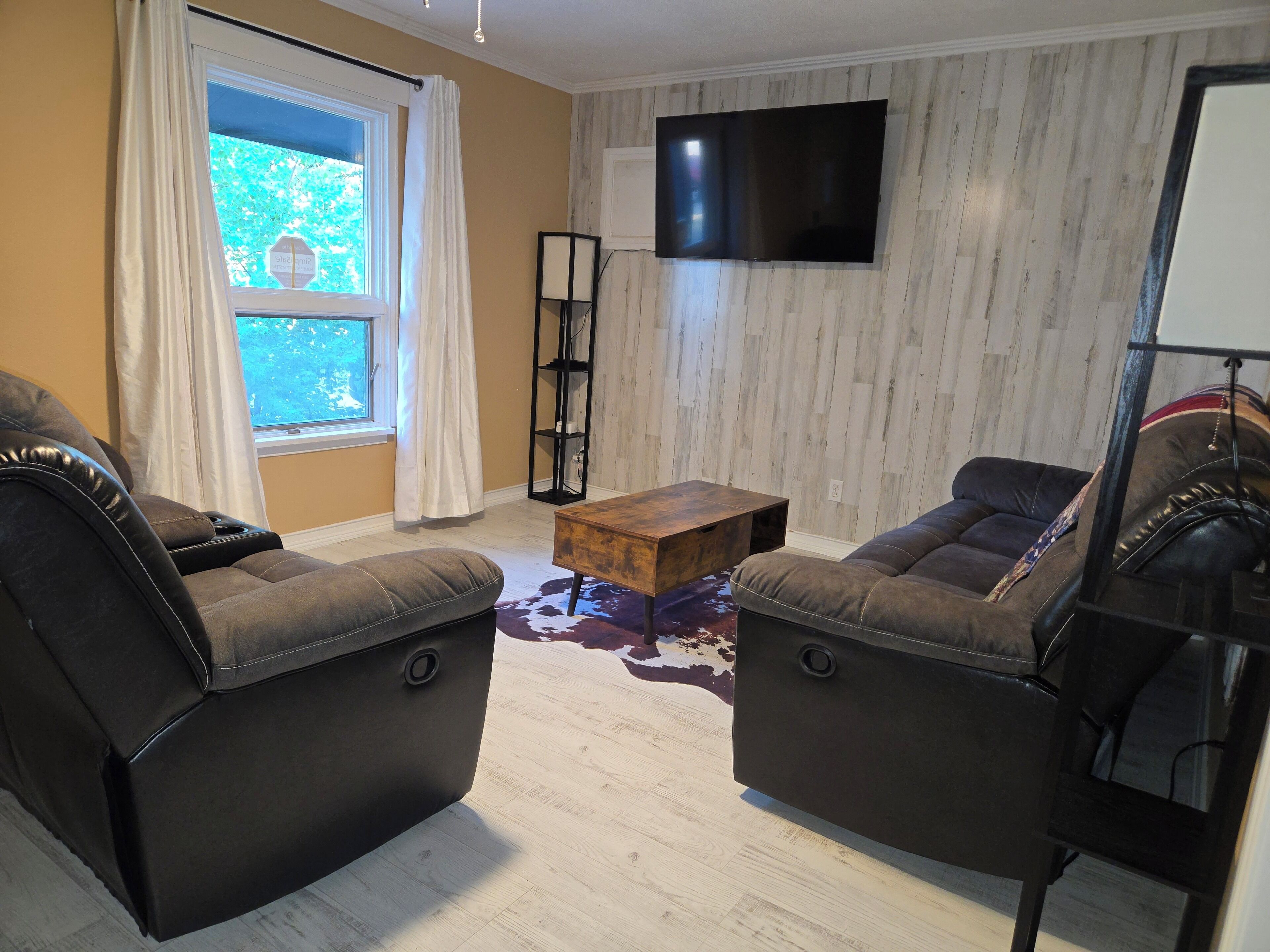 A living room with two leather sofas, a coffee table, a flat-screen TV mounted on the wall, and a window with curtains.