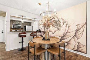 A dining area with a round wooden table, six chairs, a chandelier, and a large floral mural on the wall.