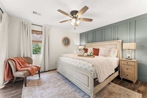 Un dormitorio con una cama, una silla, una mesita de noche, un ventilador de techo y una ventana con cortinas.