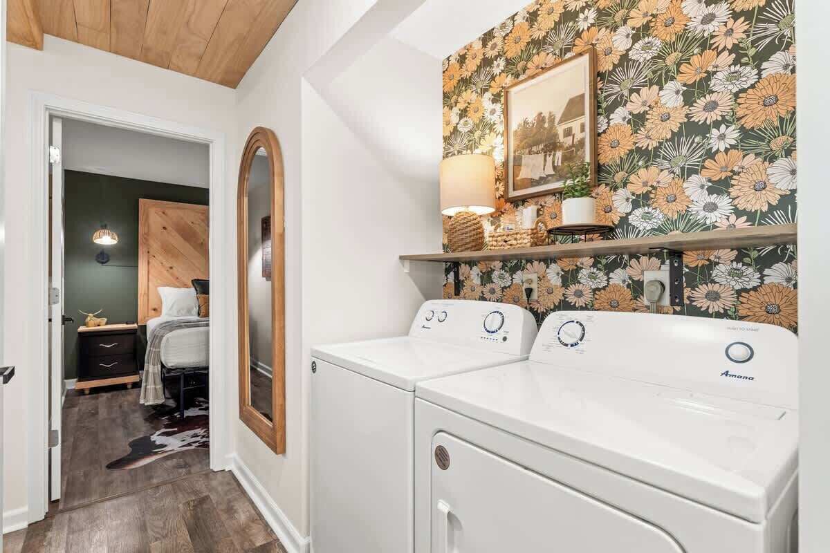 A laundry room with two white front-loading washers and dryers, a wooden door, a mirror, a wall-mounted shelf with a lamp, and a floral wallpaper.