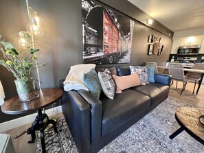 A modern living room with a black leather sofa, a round coffee table, a dining area with a chair, and a kitchen in the background.