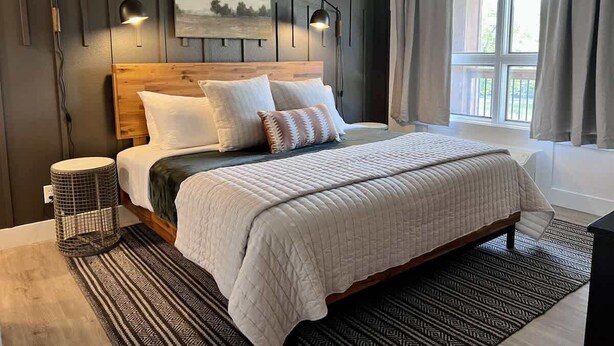 A bedroom with a wooden headboard, a large bed with multiple pillows, a bedside lamp, a floor lamp, a window with curtains, and a patterned rug. - Roark Shores by Branson Landing (Branson)