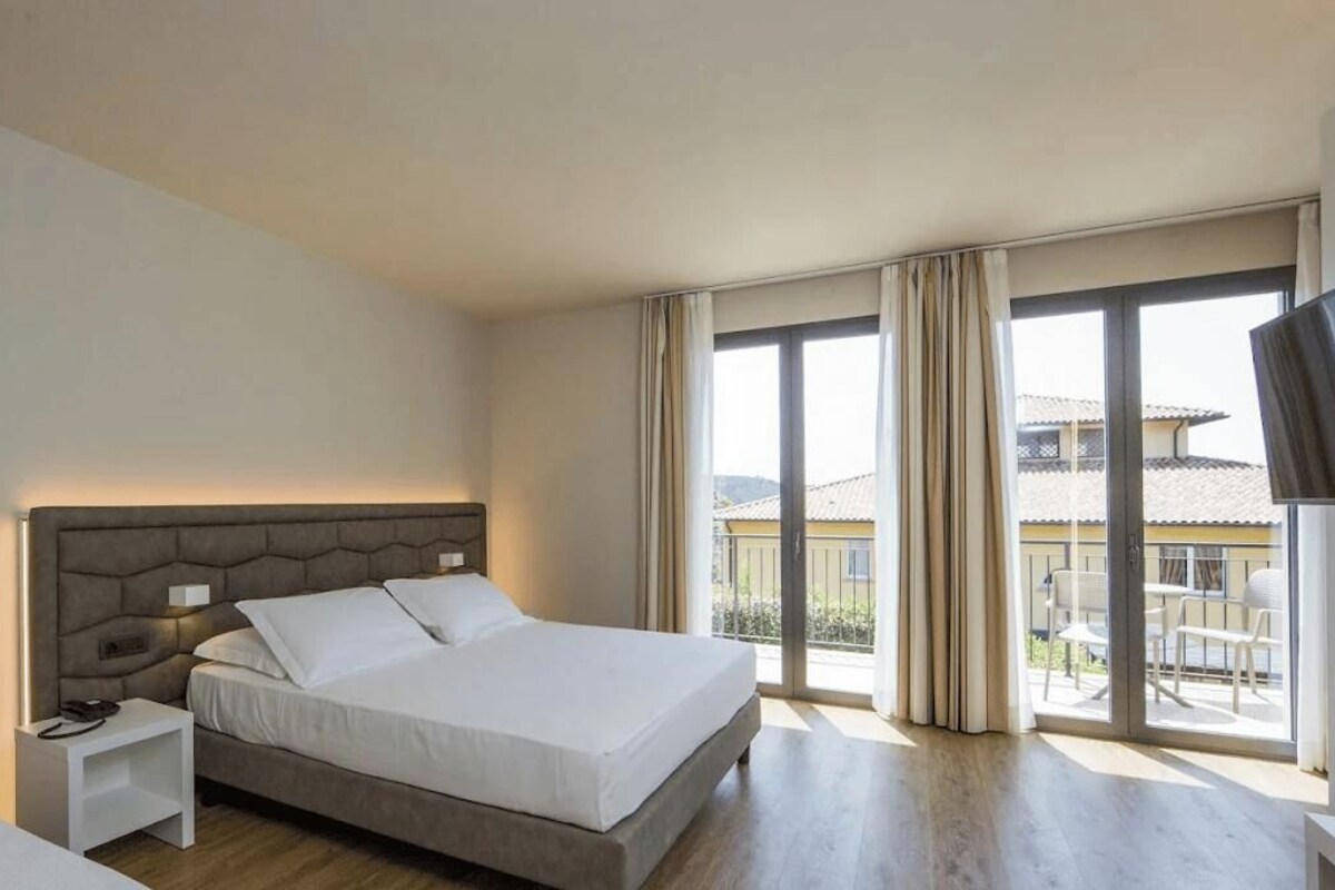 Una habitación de hotel con una cama grande, una mesita de noche, un televisor y un balcón con cortinas.