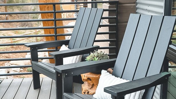 Deux chaises Adirondack noires sont posées sur une terrasse en bois, agrémentée d’une rambarde en métal. Une fenêtre offre une vue sur les montagnes, et un poteau en bois est également visible.