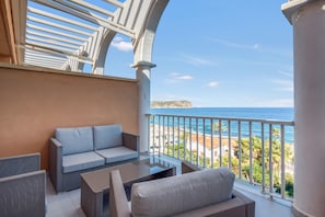 A balcony with a sofa and table, overlooking a coastal view with buildings and greenery.