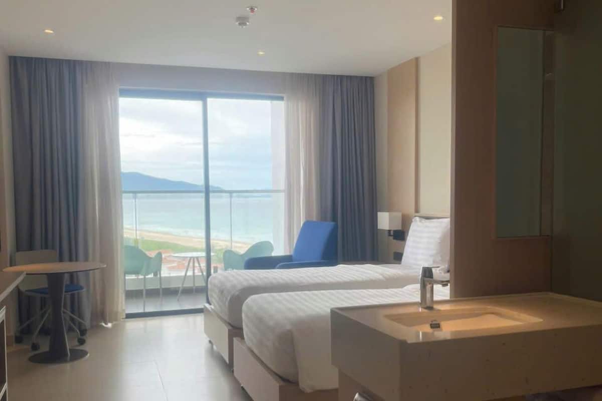 Habitación de hotel con cama, escritorio con silla, baño con lavabo y balcón con vista al mar.