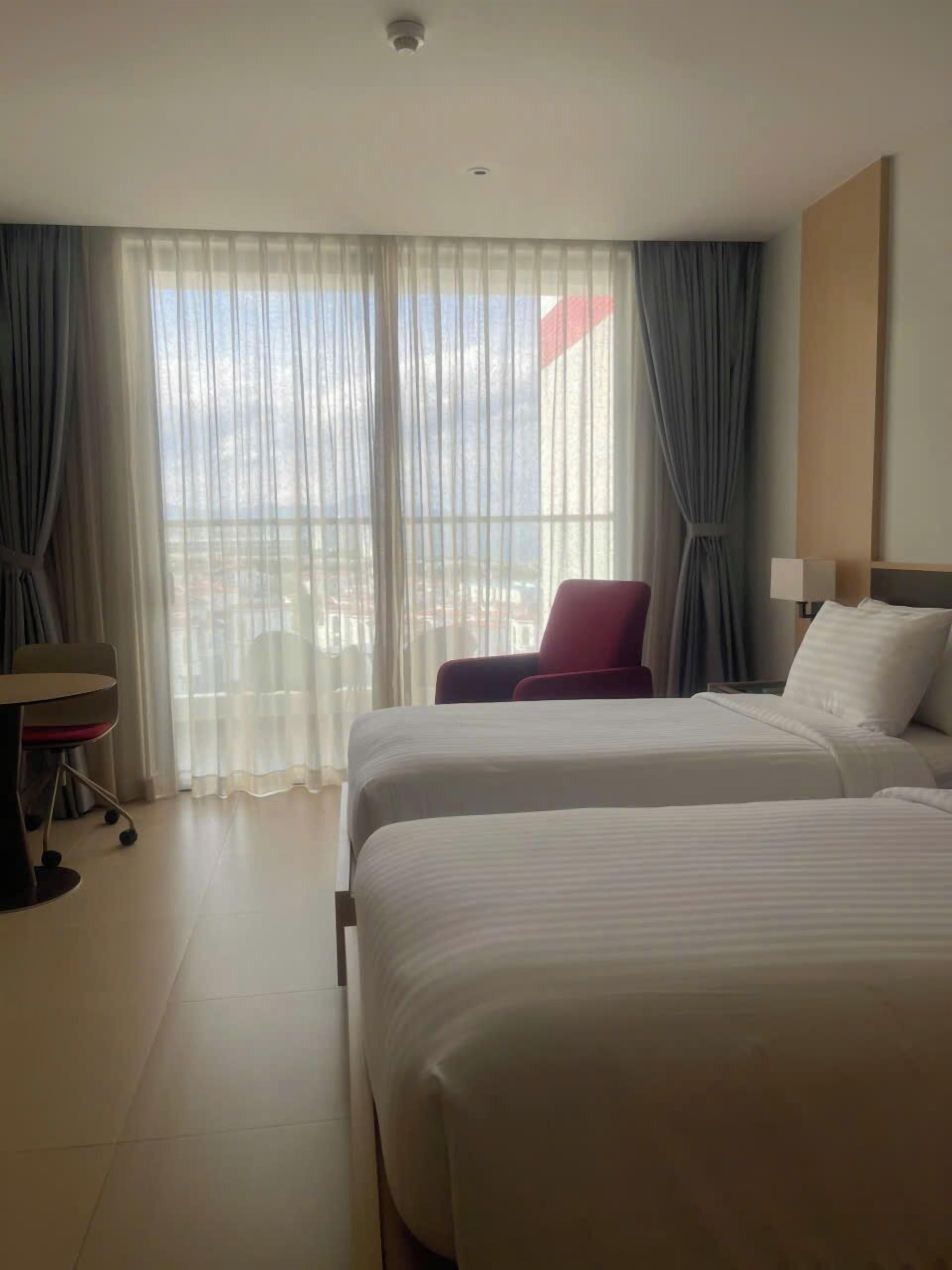 Ein Hotelzimmer mit zwei Betten, einem Schreibtisch, einem Sessel und Blick auf die Stadt durch transparente Vorhänge.