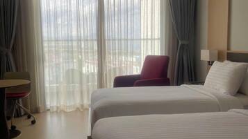 Ein Hotelzimmer mit zwei Betten, einem Schreibtisch, einem Sessel und Blick auf die Stadt durch transparente Vorhänge.