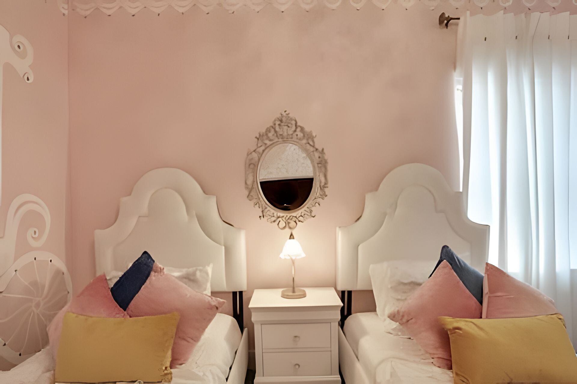 A bedroom with two beds, a nightstand with a lamp, a mirror, and a window with curtains.