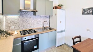 A compact kitchen with a gas stove, oven, sink, and a white refrigerator. There is a dining table with chairs and a herringbone-patterned floor. - Anna Cerasuolo House | Whole house in mountain hamlet (Molise)