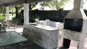 Une terrasse extérieure avec un barbecue en pierre, un espace salon et une table en verre.