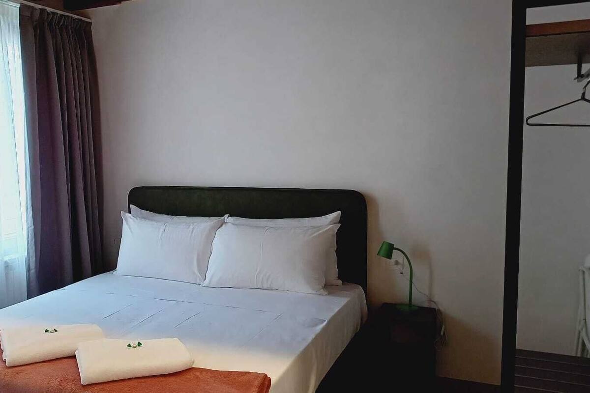 A neatly made bed with white linens and pillows, a bedside table with a lamp, and a window with curtains.