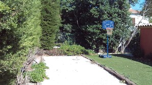 A concrete pathway leading to a basketball hoop, surrounded by greenery and a fence.