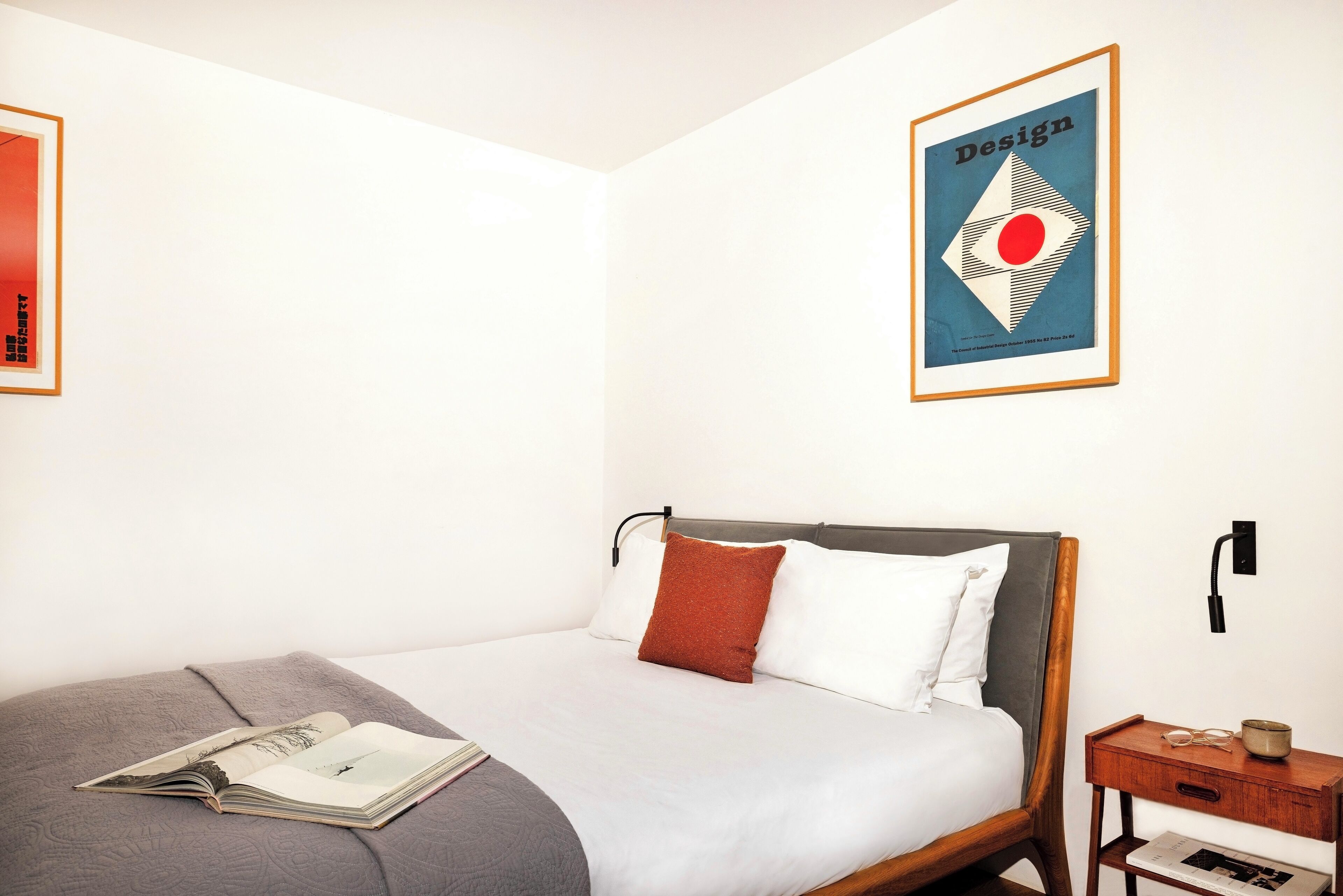 Un lit bien fait, avec une couverture grise et un oreiller rouge, une table de chevet avec une lampe, et un tableau encadré accroché au mur.