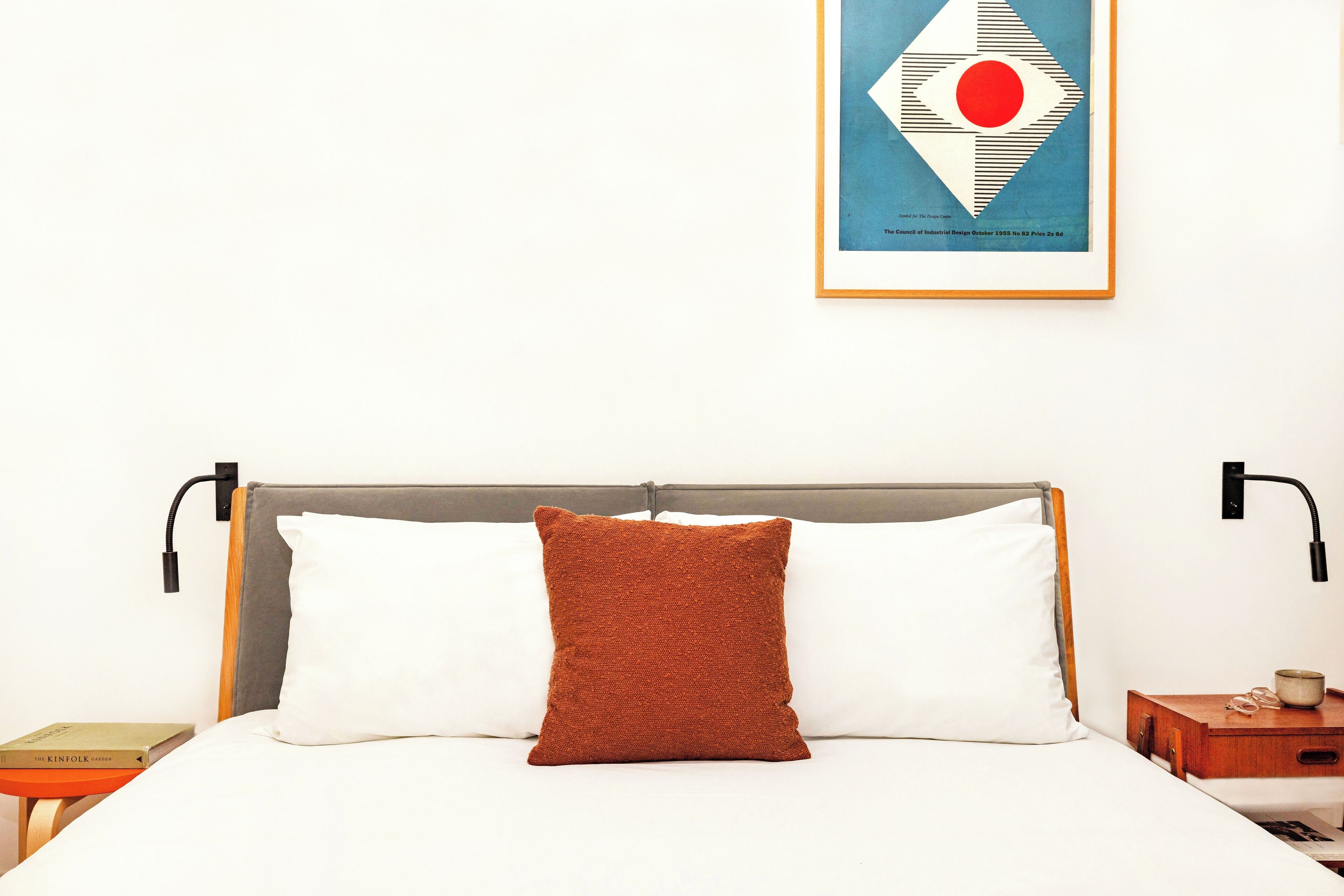 A neatly made bed with a brown pillow and white pillows, a framed picture on the wall, and a bedside table with a book and a cup.