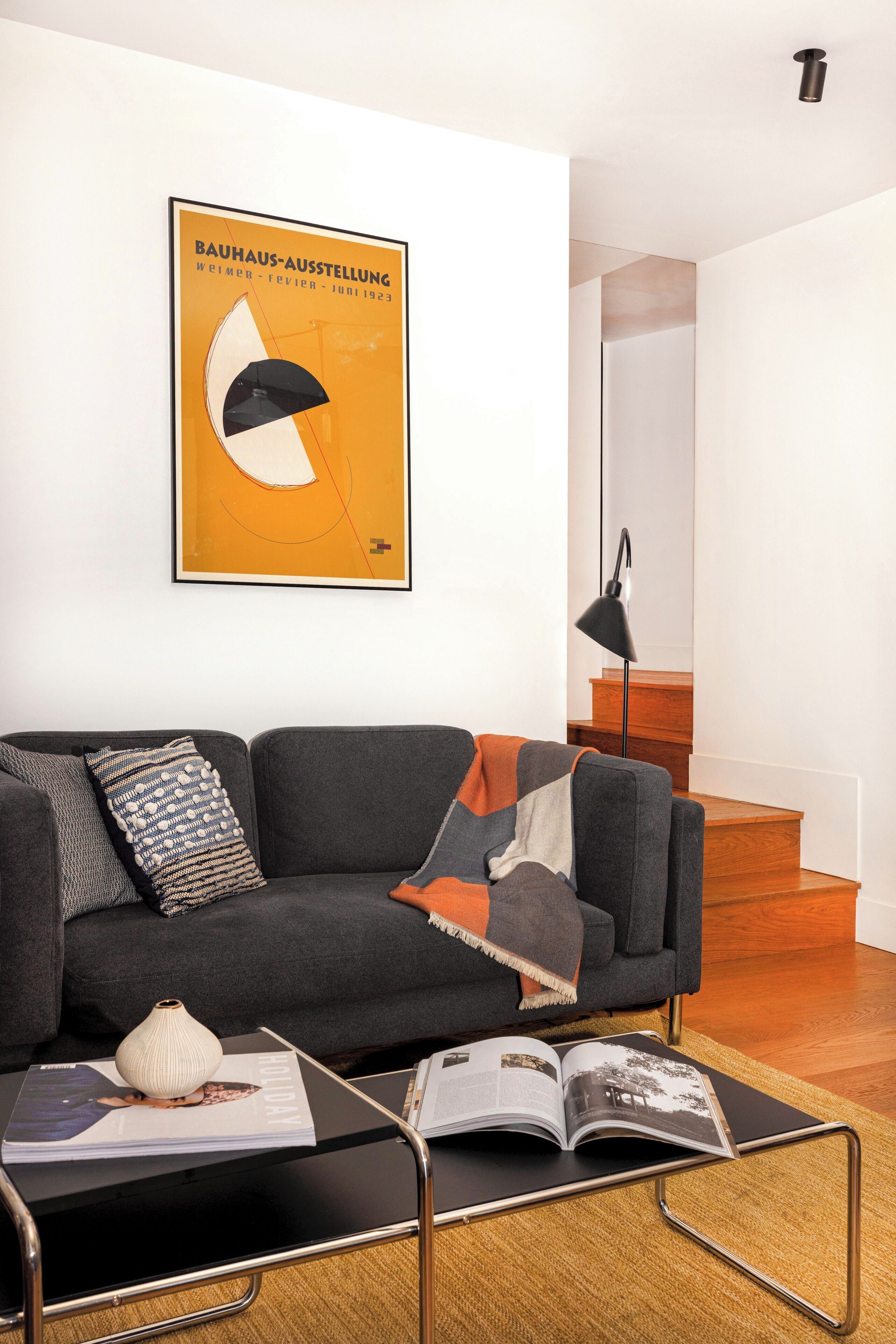 A modern living room with a grey sofa, a black coffee table with a magazine, a framed poster on the wall, and a staircase in the background.