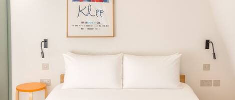 A hotel room with a bed, a small orange stool, and a colorful artwork on the wall.