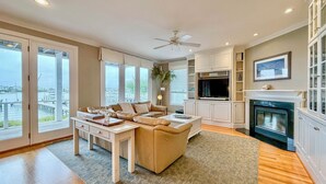 A spacious living room with a large sofa, a coffee table, and a fireplace. There is a TV mounted on the wall and a ceiling fan. - The Channel House~Water Views~Dock + Boatslip~Walk to beach! (Wrightsville Beach)