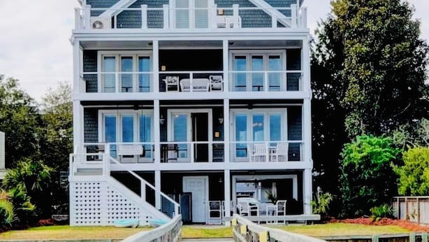 A multi-story building with a balcony on the second floor, a covered porch on the ground floor, and a wooden walkway leading to the entrance. - The Channel House~Water Views~Dock + Boatslip~Walk to beach! (Wrightsville Beach)