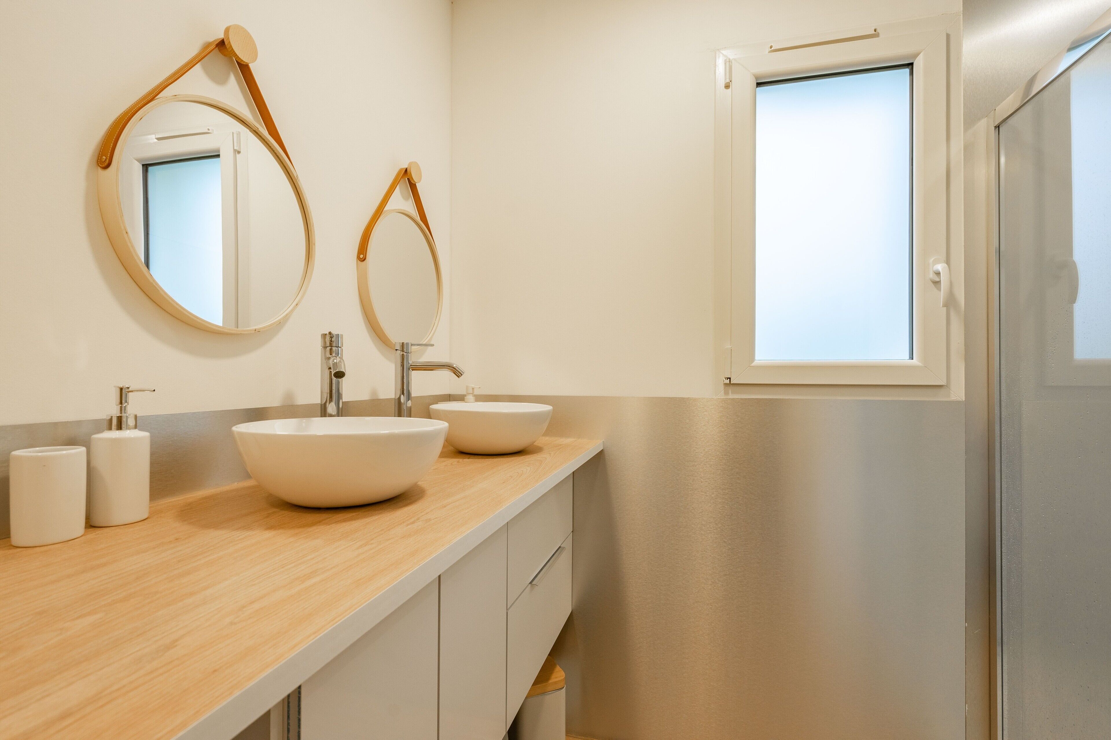 Ein modernes Badezimmer mit zwei runden Spiegeln, einer Holzauskładung, zwei Waschbecken und einem Fenster.