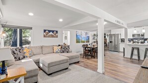 A spacious living area with a grey sectional sofa, a dining table with chairs, and a kitchen in the background. - 25 min to Yosemite - BBQ - Hot Tub -  Private Creek (Oakhurst)