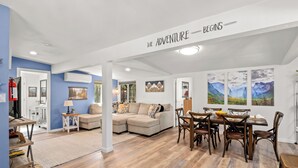 A spacious living area with a large sofa, dining table, and chairs. There is a fireplace, artwork on the walls, and a ceiling sign that reads "THE ADVENTURE BEGINS". - 25 min to Yosemite - BBQ - Hot Tub -  Private Creek (Oakhurst)