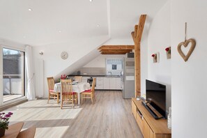 Une cuisine moderne avec des meubles blancs, un coin repas avec des chaises en bois et un téléviseur posé sur un meuble en bois.