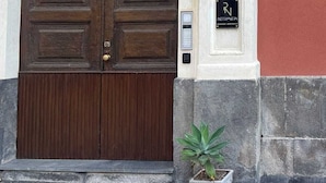 A large wooden double door with a keypad lock, a potted plant beside it, and a sign with a letter 'R' on a building facade. - Pietranera Home (Catania)