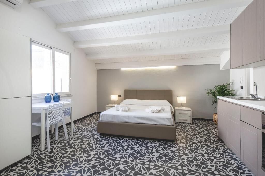 Une chambre d’hôtel comprenant un lit, des tables de chevet, un bureau avec des chaises, une fenêtre et un coin cuisine.