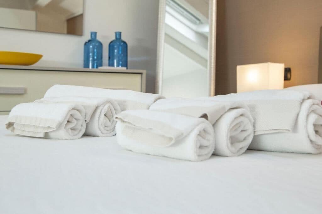 A neatly made bed with rolled white towels, a mirror, and a bedside table with a lamp and two blue bottles.