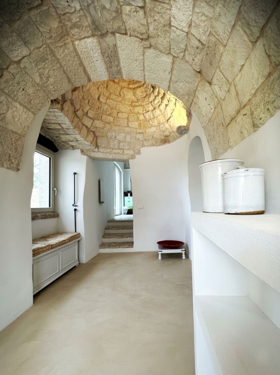 A narrow hallway with a stone ceiling, a staircase, and a white shelf with kitchenware.