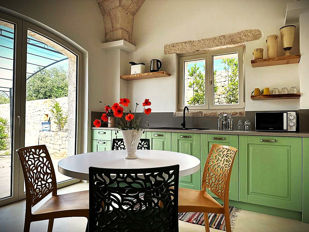 A kitchen with green cabinets, a white dining table with chairs, a vase with red flowers, and a window with a view of greenery.