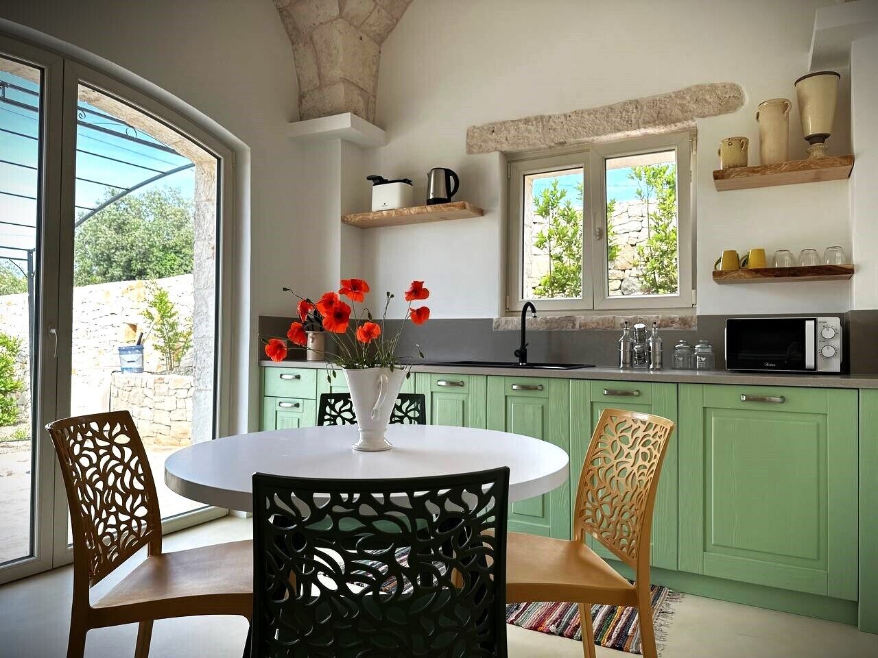 A kitchen with green cabinets, a white dining table, and chairs. There is a window with a view of greenery outside.