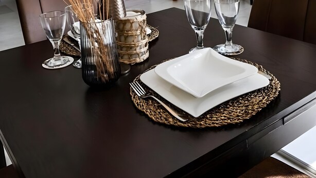 A dining table set with clear glassware, a woven placemat, and a vase with dried flowers. A white chair and a brown leather chair are positioned behind the table.