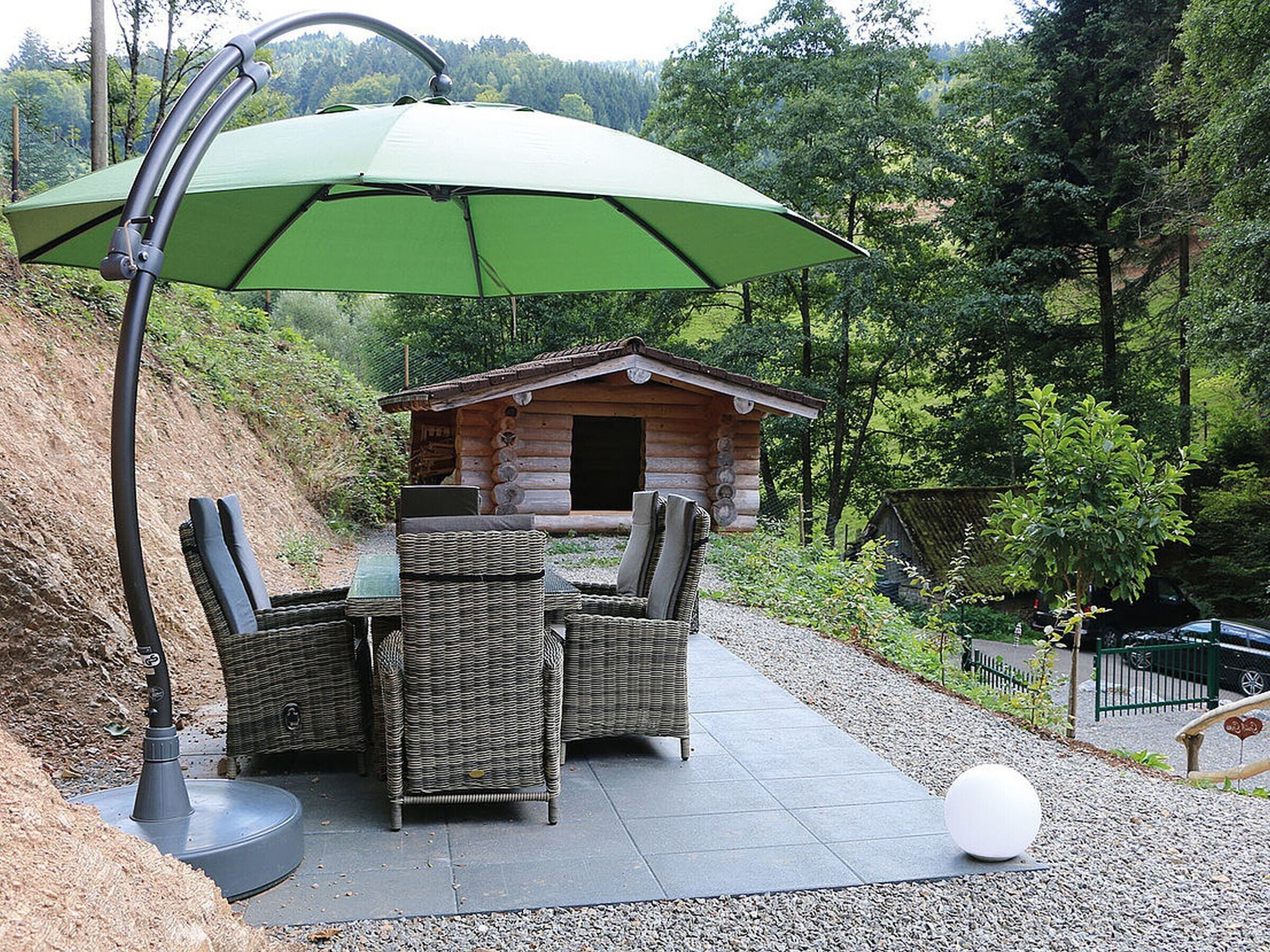 Eine Terrasse mit Korbmöbeln, einem grünen Sonnenschirm und einem Blockhaus im Hintergrund.