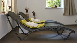 Een kamer met een ligstoel, een raam met uitzicht op groen, en een klein plankje met decoratieve voorwerpen.