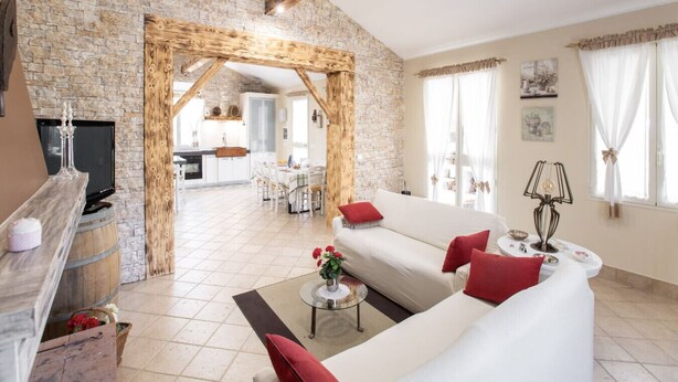 Un salon spacieux avec un mur en pierre, un canapé blanc, des coussins rouges, une table basse ornée de fleurs et un téléviseur posé sur un meuble en bois.