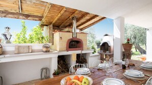 Un espace de restauration en plein air avec une table en bois rustique mise pour un repas, un sol en pierre et un four en briques en arrière-plan.