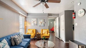 A living room with a blue sofa, two yellow armchairs, a glass coffee table, a ceiling fan, and a kitchen area with a refrigerator.