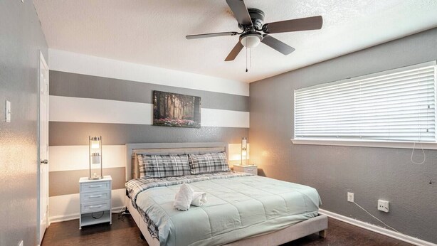 A bedroom with a bed, bedside lamps, a ceiling fan, a window with blinds, and a painting on the wall.