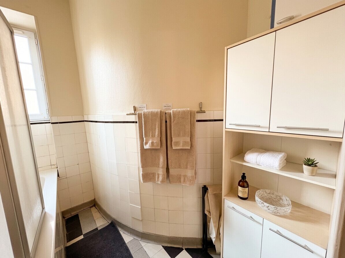 Une salle de bain carrelée de blanc, un tapis de sol à carreaux noirs et blancs, un porte-serviettes avec deux serviettes, une petite plante en pot et un meuble contenant des articles de toilette.