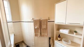 Une salle de bain carrelée de blanc, un tapis de sol à carreaux noirs et blancs, un porte-serviettes avec deux serviettes, une petite plante en pot et un meuble contenant des articles de toilette.